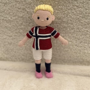 May include: A handmade crochet doll featuring blonde hair, a red shirt with a blue and white cross pattern, white shorts, dark blue socks, and pink shoes. The doll has a light skin tone, black bead eyes, and a stitched mouth on a round head.