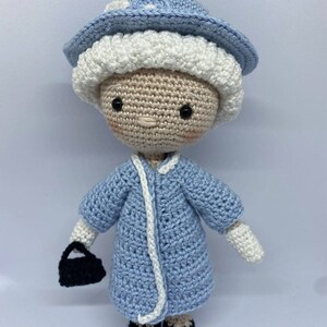 Puede incluir: Una muñeca de crochet que lleva un vestido azul, guantes blancos y un sombrero azul y blanco. La muñeca lleva un bolso negro.