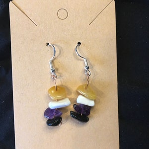 May include: A pair of dangle earrings with four stacked gemstone beads. The beads are yellow, white, purple, and black. The earrings are on a brown card.
