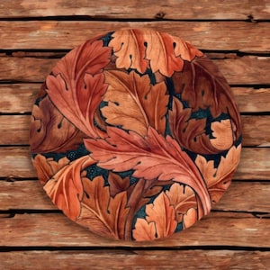 May include: Circular decorative plate with a detailed pattern of stylized leaves in shades of orange, brown, and red. The leaves are set against a dark teal background. The plate is placed on a wooden surface.