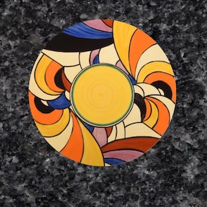 May include: A round ceramic plate with a colourful abstract design. The plate features a central yellow circle surrounded by swirling lines in shades of orange, blue, black, and purple.