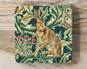 May include: Square coaster featuring a brown hare amidst a floral design with red and white flowers, set against a dark teal background. The coaster has a decorative, vintage aesthetic.