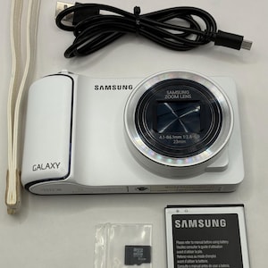 Puede incluir: Cámara digital Samsung Galaxy blanca con lente de zoom, cable de carga negro, batería, tarjeta de memoria y correa para la muñeca. La cámara tiene la palabra "GALAXY" impresa.
