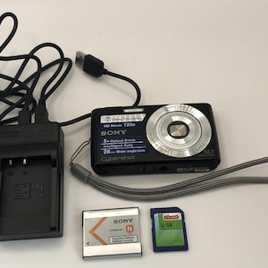 May include: A black Sony Cyber-shot digital camera with a silver lens and a 5x optical zoom. The camera is accompanied by a black charger, a black memory card, and a grey memory card.