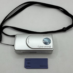 May include: A silver Sony Cyber-shot U digital camera with a black neck strap and a blue 64MB Memory Stick. The camera has a lens and several buttons. The Memory Stick is rectangular.