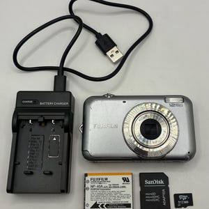 May include: A silver Fujifilm digital camera with a charger, battery, and memory cards. The camera has a lens and the text "12 Mega Pixels". The charger is black and has the text "CHARGE BATTERY CHARGER". The battery is labeled "FUJIFILM NP-45A".