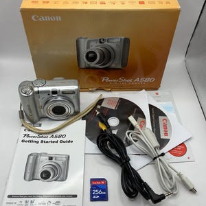 May include: A silver Canon PowerShot A580 digital camera with a strap, box, and accessories. The box is tan with the Canon logo. Included are a CD, cables, a 256MB SD card, and a user guide. The camera has a lens and buttons.