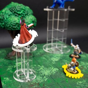 Puede incluir: Una figura en miniatura de una maga con una túnica roja y un libro en la mano, de pie sobre una base de acrílico transparente con un anillo de plástico blanco. La figura está sobre una alfombra de juego verde con un árbol y otras figuras en el fondo.