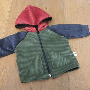 May include: A green corduroy jacket with a red hooded top and black sleeves. The jacket has a zipper closure and two pockets.