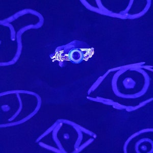 May include: A silver ring with a blue evil eye charm. The ring is surrounded by several blue glowing eyes drawn on a dark blue surface.