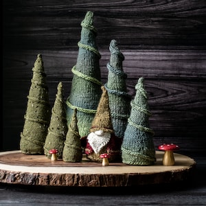 Puede incluir: Una figura de gnomo rodeada de árboles de punto verde y azul sobre una rodaja de madera. El gnomo lleva un gorro rojo y blanco y una barba blanca.
