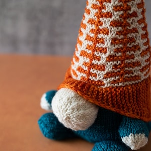 May include: A knitted gnome with a white and orange patterned hat. The gnome is sitting on a wooden surface.