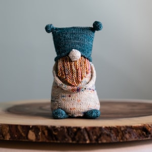 Könnte beinhalten: Ein gestrickter Zwerg mit einem türkisfarbenen Hut und einem weißen und braunen Bart. Der Zwerg sitzt auf einer Holzscheibe.