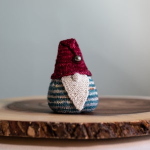 Pode incluir: Um gnomo de tricô com um chapéu vermelho e uma barba branca. O gnomo está sentado em uma fatia de madeira.