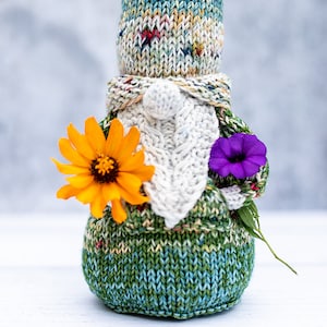 Könnte beinhalten: Eine gestrickte Gnom-Puppe mit einem weißen gestrickten Bart und einer grün-blau-weißen gestrickten Mütze. Der Gnom hält eine gelbe Blume und eine violette Blume.