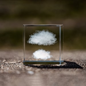 May include: A clear glass cube containing two white clouds. The cube is resting on a rough, brown surface.