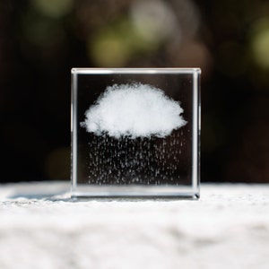 Op de afbeelding: Een heldere glazen kubus met een witte wolk en gegraveerde regendruppels erin. De kubus staat op een wit oppervlak.