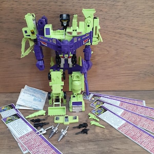 May include: A lime green and purple robot action figure with a black head, accompanied by several small plastic accessories, including black guns, silver drill bits, and light green components, along with printed paper instruction manuals.