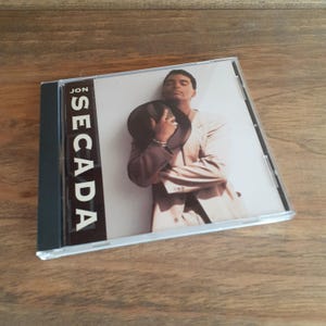 May include: A Jon Secada music CD in a clear plastic jewel case. The cover art features a photograph of a person in a light-colored blazer holding a dark hat against their chest. The text JON SECADA is printed vertically in white on a black bar.