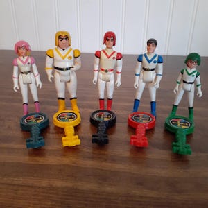 May include: Five vintage action figures with matching key-shaped accessories. The figures are dressed in white, with accents of pink, yellow, red, blue, and green. Each figure stands on a key with a circular top and a cross design.