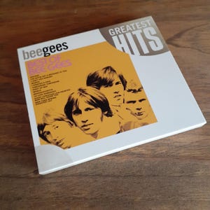 May include: A square CD case for the Bee Gees Greatest Hits album. The cover features a white background with a large yellow section displaying a black and white photograph of the band members. The text reads Bee Gees, Best of Bee Gees, and Greatest Hits, along with a tracklist including Holiday, Words, and Massachusetts.
