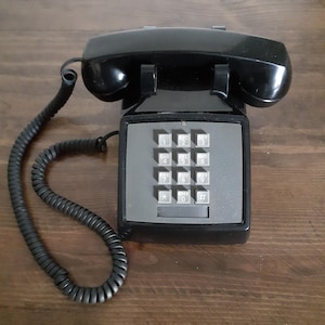 May include: Black, vintage telephone with a gray keypad and a coiled handset cord. The keypad features numbered buttons and a rectangular design. The phone is set on a wooden surface.
