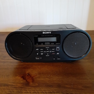 May include: A black Sony portable radio with a CD player, USB port, and Bluetooth capabilities. The radio has two large speaker grilles on either side of a digital display and control buttons. The unit is on a wooden surface.