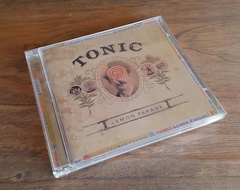May include: A Tonic Lemon Parade music CD in a clear plastic jewel case. The cover features a sepia-toned vintage portrait of a person with a red target over their eye, surrounded by decorative botanical illustrations and the text Tonic Lemon Parade.