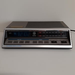 May include: A General Electric digital alarm clock radio with a silver and dark gray finish. The front panel features a digital time display, an analog AM/FM radio tuner, and various buttons labeled Time Set, Radio, and 2 Wake Times.