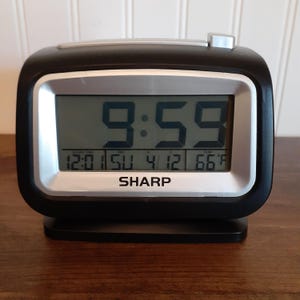 May include: A black digital alarm clock with a silver frame displays the time as 9:59. The clock also shows the alarm time, date, and temperature in Fahrenheit. The brand name "SHARP" is printed at the bottom.