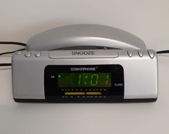 May include: A silver Conairphone alarm clock radio with a black digital display showing 1:01. The device features a prominent snooze button on top, AM and alarm indicators, and a radio frequency tuner scale with a yellow needle indicator.
