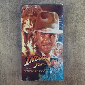 May include: A vintage VHS tape of "Indiana Jones and the Temple of Doom." The cover features a portrait of Harrison Ford as Indiana Jones, with other characters and fiery imagery. The title is prominently displayed in a stylized font.