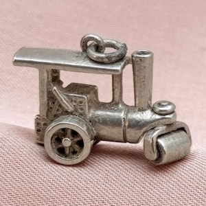 May include: A small, silver-colored charm in the shape of a steamroller. The charm has a detailed design, including wheels, a roller, and a smokestack. The charm is likely made of metal and is designed to be attached to a bracelet or necklace.