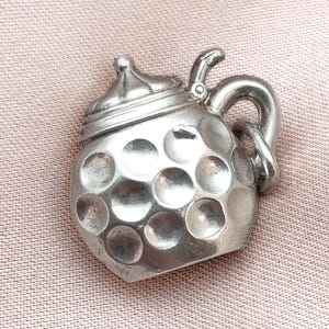 Puede incluir: Broche plateado con forma de jarra de cerveza, con tapa y asa. El cuerpo de la jarra tiene una textura con hoyuelos. La broche está sobre un fondo rosa.