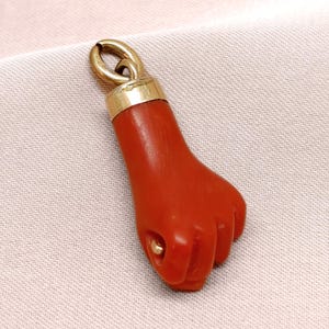 May include: A red coral fist pendant with a gold-colored bail. The pendant is a detailed carving of a clenched fist, showcasing the knuckles and the shape of the hand. The bail is a simple loop for attaching to a chain.