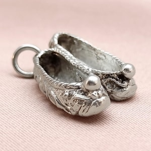 May include: A pair of miniature silver-coloured shoe charms. Each shoe features intricate detailing, a rounded toe, and a small ball at the front. A circular loop is attached to one shoe for hanging.