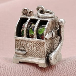 May include: A silver-toned slot machine charm with the word "JACKPOT" engraved on the front. The slot machine has three visible reels with colorful designs. A pull lever is on the side. The charm is designed to be attached to a bracelet or necklace.