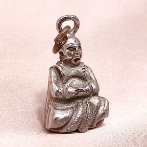 Puede incluir: Un pequeño dije plateado que representa una figura sentada con las manos juntas. El dije tiene un lazo en la parte superior para sujetarlo. La figura lleva una túnica y tiene rasgos faciales detallados. El dije es probablemente de metal.