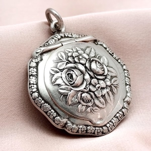 Puede incluir: Un relicario antiguo plateado con un diseño floral. El relicario presenta un relieve detallado de rosas y otras flores en la parte delantera. Los bordes están adornados con pequeños elementos decorativos. Tiene un lazo para una cadena.
