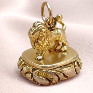 May include: A gold pendant with a lion figure on a heart-shaped base. The pendant has a loop for a chain and is set against a pink background. The base is decorated with floral patterns. The pendant is likely made of gold.