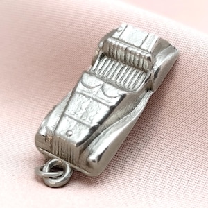 May include: A silver-colored vintage car charm with detailed engine and body features. The charm has a loop for attaching to a necklace or bracelet. The car is set against a pink fabric background.