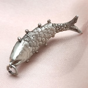 May include: A detailed silver fish charm with textured scales, fins, and a small loop at the mouth. The charm has small, round embellishments along its back and is set against a soft pink background.