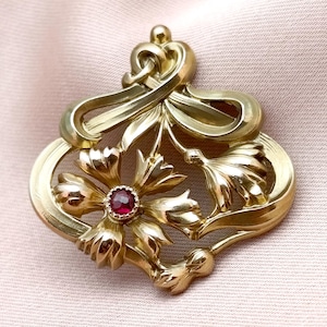 Könnte beinhalten: Eine goldfarbene Brosche mit einem stilisierten Blumendesign aus geschwungenen, bandartigen Metallelementen. In der Mitte der Hauptblüte befindet sich ein kleiner, runder, dunkelroter Edelstein. Das Metall hat eine polierte Oberfläche.