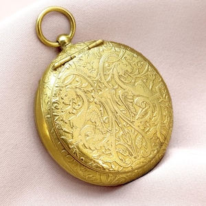 May include: A gold-colored, round antique pocket watch with intricate floral and foliate engravings. The watch has a hinged lid and a small ring at the top for attaching a chain. The watch is made of metal and has a polished finish.