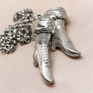 May include: A silver-toned necklace featuring a pendant of two antique-style boots. The boots are detailed with laces and textured tops, connected to a chain necklace. The boots are approximately 1.5 inches in height.
