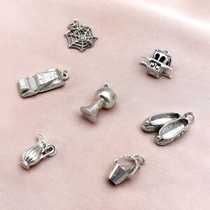 May include: A collection of silver-toned charms, including a spiderweb, a car, a goblet, a carriage, a pair of shoes, a pitcher, and a bucket. These small, detailed charms are likely for jewelry making or crafting.