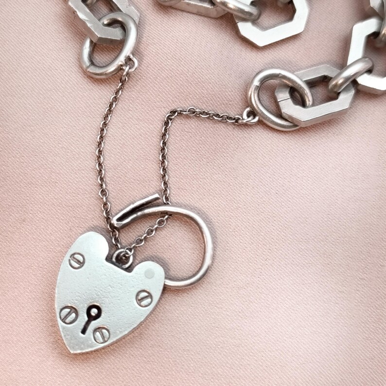 May include: A silver-toned necklace featuring a heart-shaped padlock pendant with a keyhole and decorative screws. The pendant hangs from a delicate chain, connected to a larger chain with geometric links. The background is a soft pink.