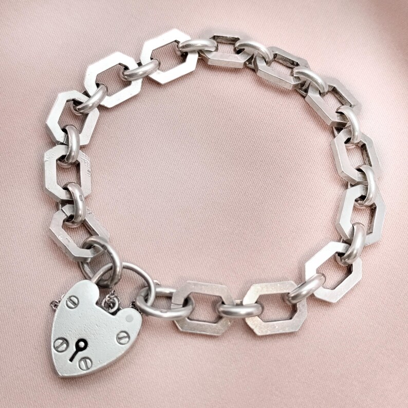 May include: Silver-toned bracelet featuring a chain-link design with geometric links. A heart-shaped padlock charm with a keyhole detail hangs from the bracelet. The bracelet is displayed on a pink surface.
