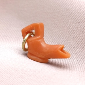 May include: A small, orange-colored boot charm with a gold-colored ring for attachment. The boot has a detailed design and is likely made of plastic or resin. The charm is a miniature representation of a cowboy boot.
