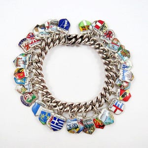 May include: A silver charm bracelet with numerous enameled shield-shaped charms. The charms feature various city names and designs, including "KOLN", "ATHEN", and "COVENTRY". The bracelet is on a white background.